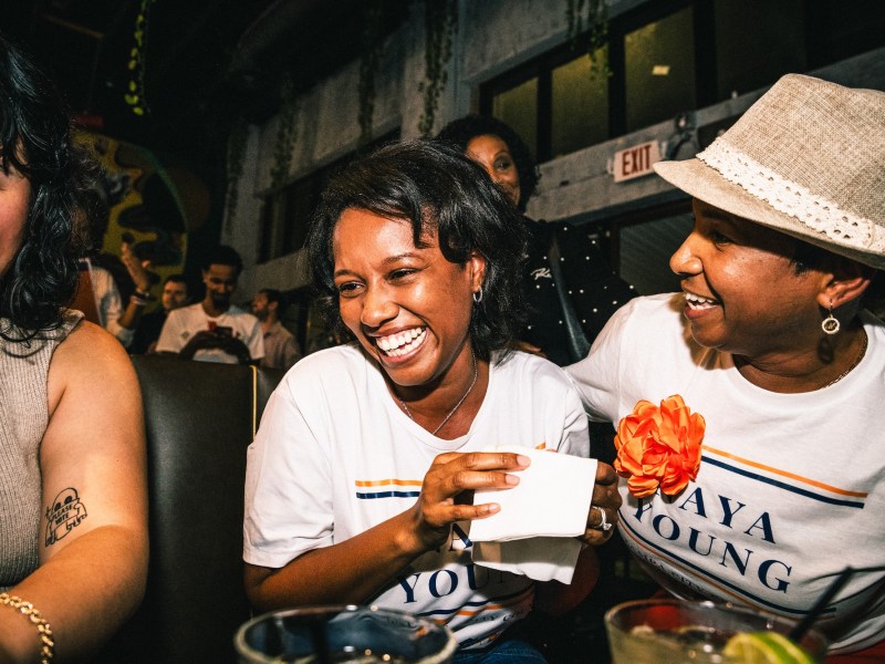 Naya Young (center) smiles and laughs with supporters at a victory party, holding a napkin. She and the woman to her right are wearing white T-shirts that read "NAYA YOUNG" in orange and blue text.