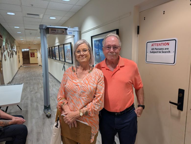 A man and a woman stand side-by-side in a building hallway, smiling for the camera. The woman on the left has gray hair and wears an orange and white patterned shirt. The man on the right has a mustache and wears an orange polo shirt. In the background, a walk-through metal detector is to their left, and a sign on a door to their right reads, "ATTENTION All Persons are Subject to Search."