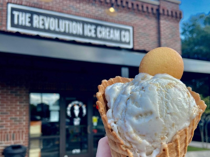 A close-up shot features a waffle cone filled with a large scoop of vanilla ice cream speckled with crumbs and topped with a round vanilla wafer cookie. The treat is held up against a blurred background showing the brick storefront and sign of "The Revolution Ice Cream Co".