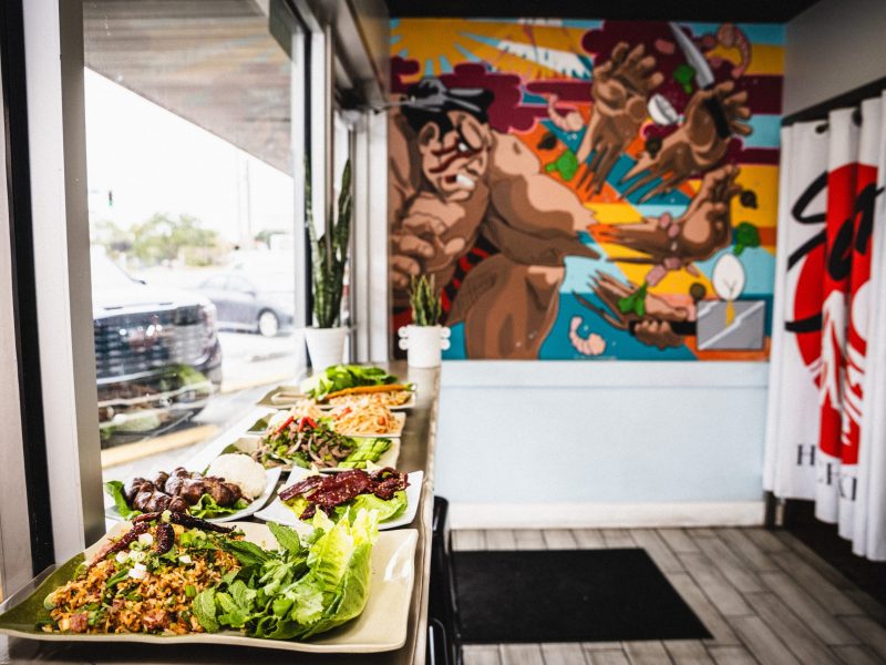 Interior view of the restaurant near the window, showing multiple plates of various Laotian dishes, including salads, meats, and rice, lined up on the counter in front of the mural.