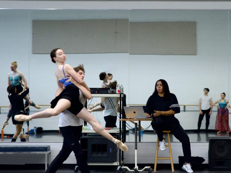 A choreographer or director sitting on a stool to the right, looking at dancers performing a lift in a brightly lit studio with a mirrored wall. The woman is wearing a black top and pants, working on a silver laptop resting on a cart. Other dancers are visible in the background and reflections.