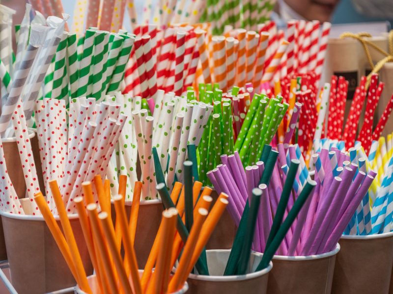 A close-up display of assorted colorful paper straws arranged in small brown cups. The straws feature a variety of vibrant patterns, including diagonal stripes and polka dots in red, green, blue, yellow, and purple, alongside solid-colored orange and purple straws.
