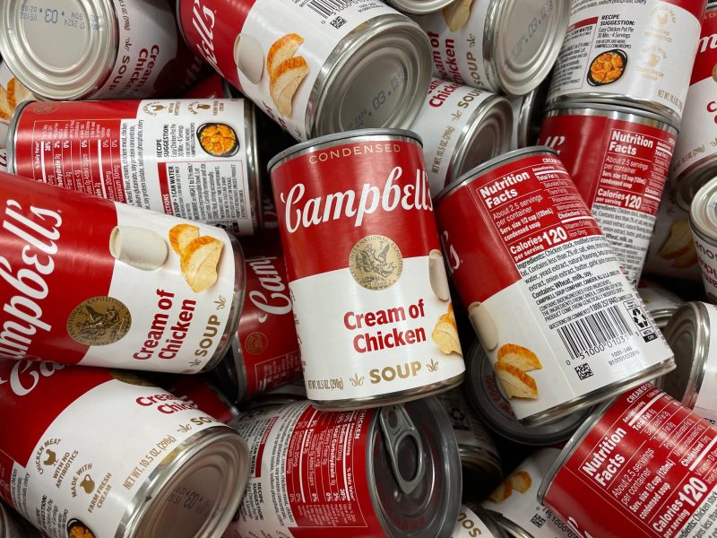 A disorganized pile of Campbell's Cream of Chicken soup cans fills the frame. The classic red and white labels are visible from various angles, displaying the brand logo, product name, and nutrition information tables.