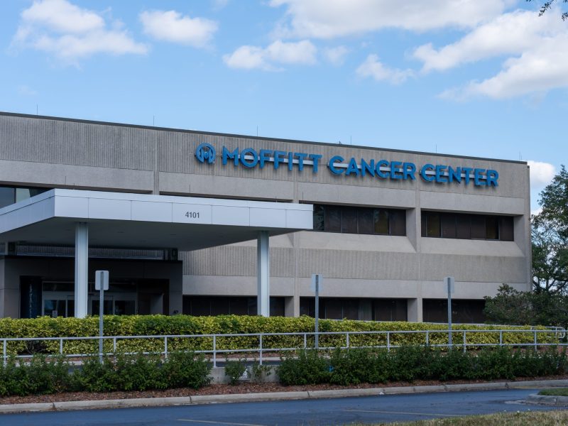 A daytime exterior view of the Moffitt Cancer Center. The large, beige concrete building features a covered entrance canopy with the number "4101" visible. Prominent blue lettering on the upper façade reads "MOFFITT CANCER CENTER." Green hedges and landscaping line the front of the building under a bright blue sky with scattered clouds.