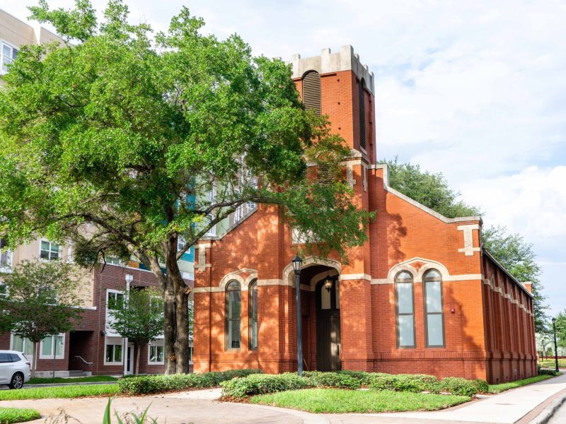 The historic St. James Church, a red brick structure, is seen on a sunny day in downtown Tampa, situated on a grassy corner lot adjacent to a new apartment complex and a paved sidewalk.