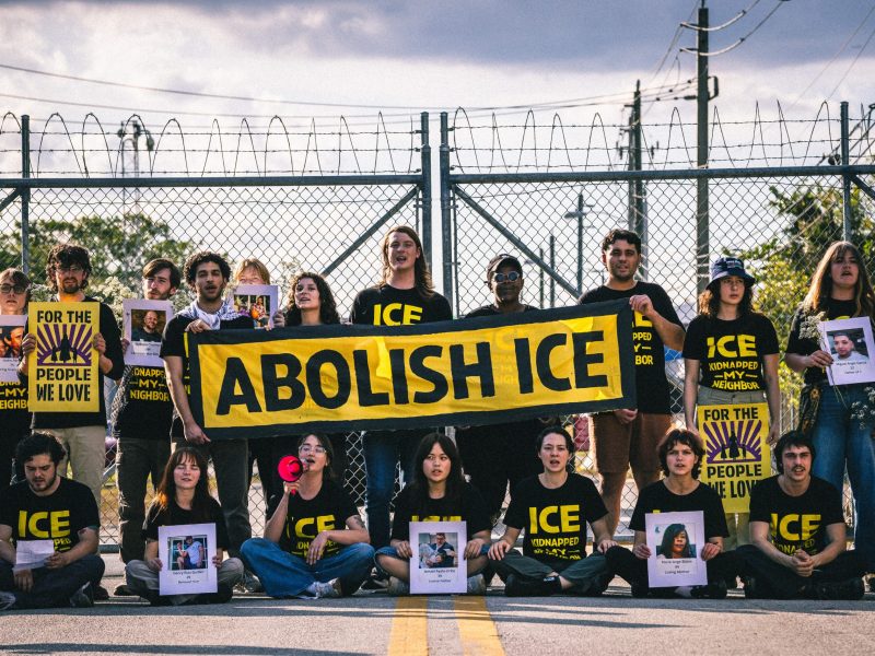 A group of protestors stands and sits in front of a chain-link fence topped with barbed wire, holding a large yellow banner that reads "ABOLISH ICE." Many wear black shirts and hold signs with photos of people.