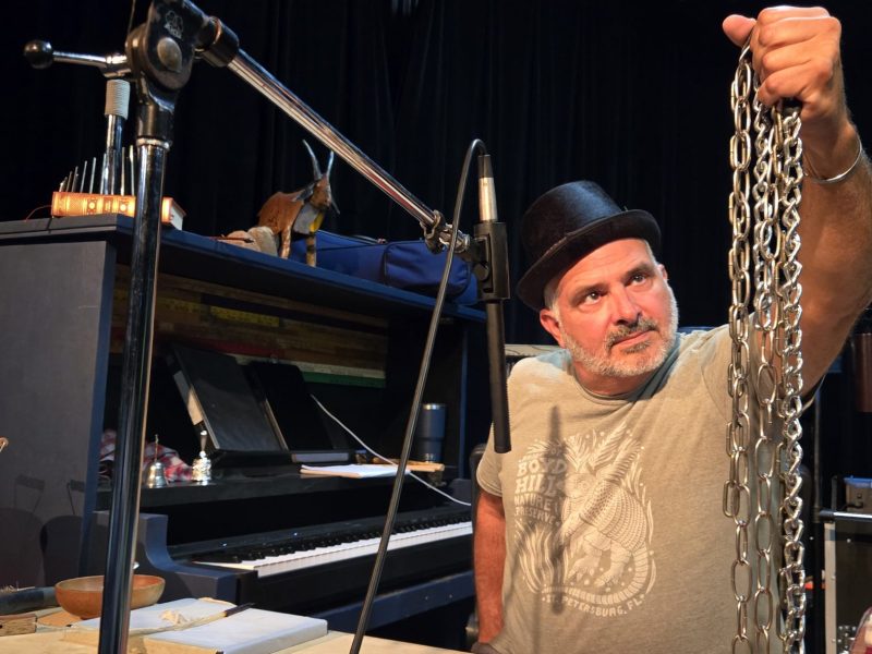An individual, identified as Juan Rodriguez, performing as a Foley artist on stage. The person, wearing a fedora and a graphic t-shirt, holds up a heavy metal chain in the air while looking towards it. A piano, a microphone, and various sound props are set up around them.