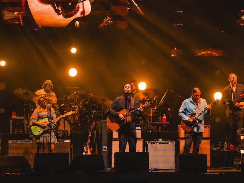 The large Tedeschi Trucks Band performs on stage under warm amber spotlights with a giant projection of an acoustic guitar above them.