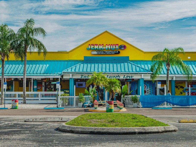 Exterior view of a bright yellow and turquoise restaurant building named "JERK HUT," with tall palm trees lining the front patio and a blue sky overhead.