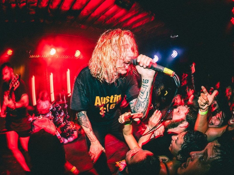 The lead singer of a band with long blonde hair and heavy arm tattoos leans over the stage, screaming into a microphone toward the audience. The crowd is pushing against the stage barrier, raising their hands and cheering. The stage is bathed in harsh red stage lighting.