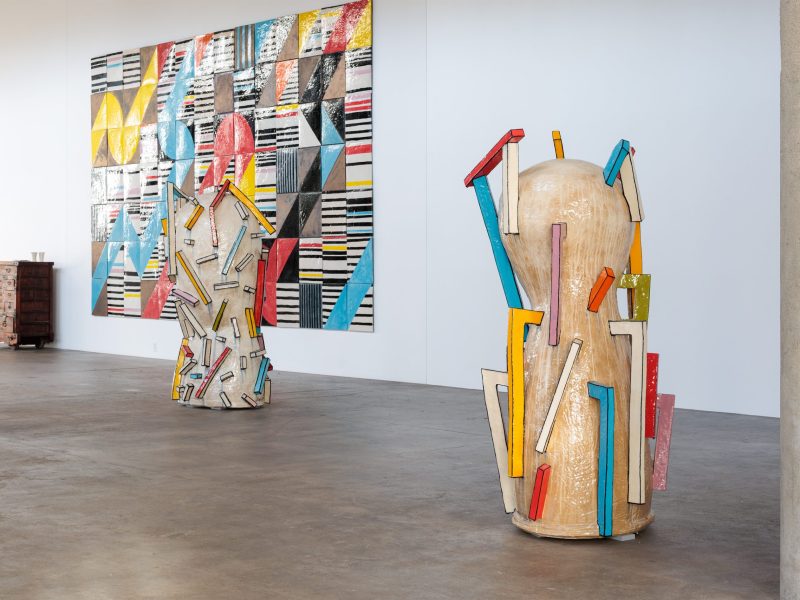 Two tall, abstract, beige sculptures with colorful geometric protrusions stand on a concrete floor in front of a large, colorful, tiled geometric wall artwork in a bright gallery space.