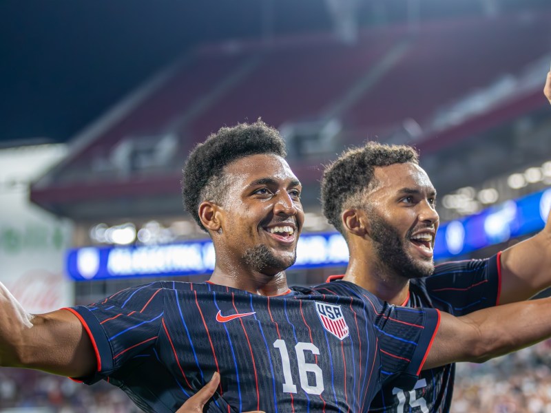 Two U.S. Men's National Team (USMNT) soccer players, one wearing jersey number 16, smile and raise their arms in celebration on the field after a goal or victory, with the stadium stands visible in the background.