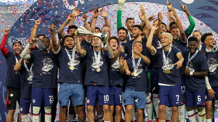 U.S. Men’s National Soccer Team players celebrate on stage with medals and confetti after winning a championship, holding up the trophy and cheering together.