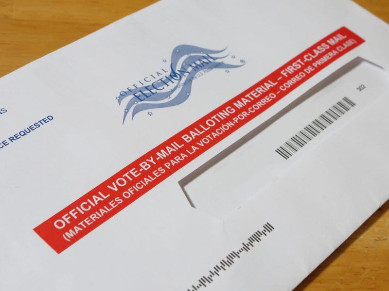 Close-up of a white Official Election Mail envelope with a red stripe reading "OFFICIAL VOTE-BY-MAIL BALLOTING MATERIAL" in English and Spanish across the center.
