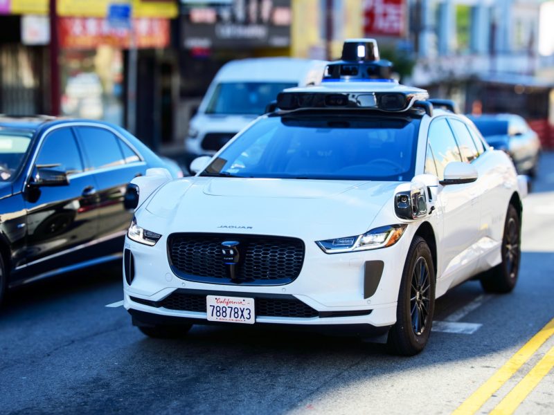 A self-driving white Jaguar I-PACE with a California license plate is parked or driving in a sunny city street, with other cars and storefronts blurred in the background. The vehicle's distinctive black Waymo sensor hardware is clearly visible on the roof.