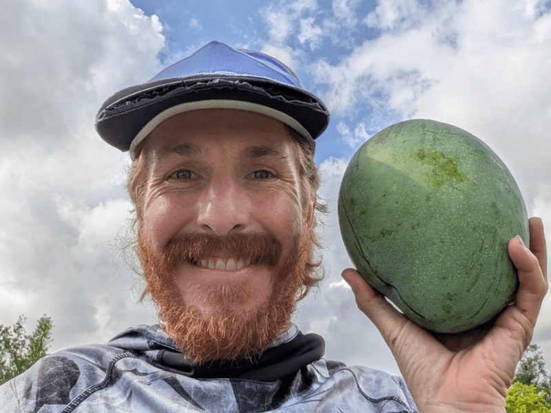 Smiling man with a red beard and mustache wears a patterned shirt and sun visor, holding a large green fruit up to the camera. A partly cloudy sky fills the background.