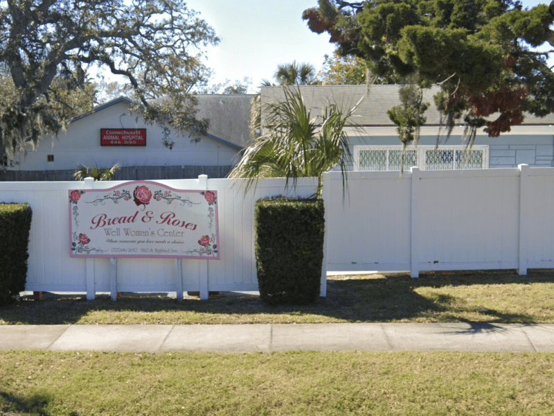 A daytime street view of a small community building behind a white fence and trimmed hedges. The sign is visible in the foreground, with the single-story clinic building and surrounding residential trees in the background.
