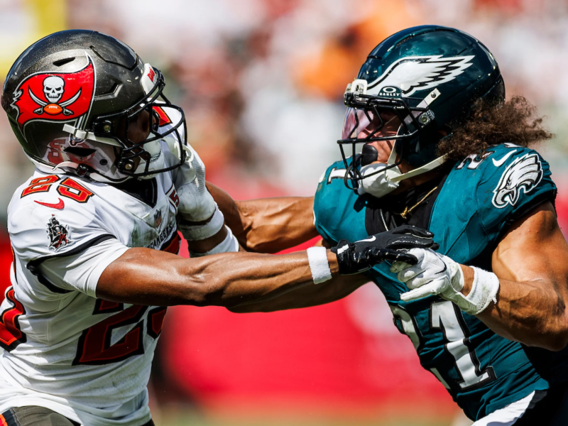 Two football players are engaged in a play on the field. A player from the Tampa Bay Buccaneers in a white jersey with the number 25 is pushing off a player from the Philadelphia Eagles, who is wearing a teal jersey with the number 11 and a winged helmet.