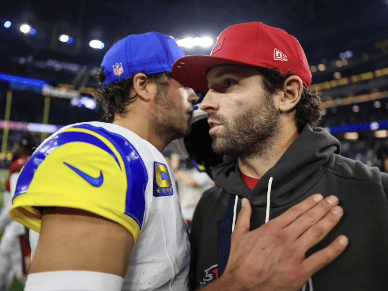 Close-up of two football players talking intensely on the field after a game; one wears a Rams uniform with a blue cap and the other wears a red cap and gray hoodie, with their hands resting on each other's chests.