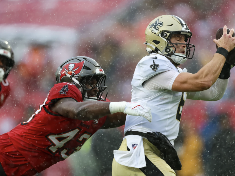 A New Orleans Saints player wearing a white jersey with the number 9 is focused on catching the football while a Tampa Bay Buccaneers player in a red jersey attempts a tackle from behind, pulling at the back of the white jersey.