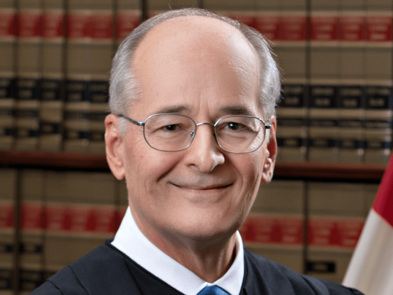 Official portrait of Florida Supreme Court Justice Charles Canady, smiling and wearing glasses and judicial robes, against a backdrop of law books.