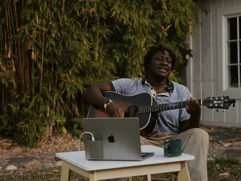 Musician Dylan Dames plays an acoustic guitar and sings outdoors, seated in front of a white building and dense bamboo plants. A laptop sits on a small table in front of him.