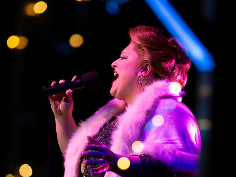 A performer with dark hair styled up and red lipstick sings with an open mouth, holding a black microphone. Warm bokeh lights and a bright blue vertical beam of light are visible in the dark background.