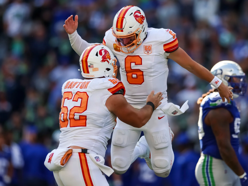 Quarterback Baker Mayfield (number 6) jumps up high in celebration, with his teammate Graham Barton (number 62) supporting him from below. Both are wearing the Tampa Bay Buccaneers' white and orange throwback uniforms on the field.