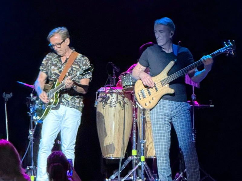 Two musicians perform on stage under bright lights. One plays an electric guitar, wearing glasses, a patterned shirt, and white pants. The other plays a bass guitar, dressed in a dark shirt and checkered pants. Behind them are conga drums and other percussion instruments, with stage lighting adding purple and blue highlights. Audience members are faintly visible in the foreground.