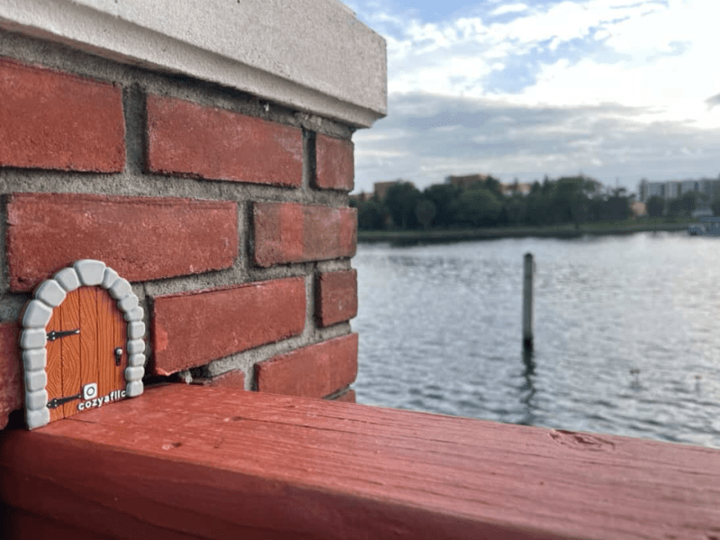 A miniature, arched wooden door with gray stone trim is affixed to the corner of a red brick column, resting on a reddish-brown wooden railing. The door has a small white tag at its base with "cozyafllc" and an Instagram logo. In the background, a body of water stretches out with a single wooden piling visible.