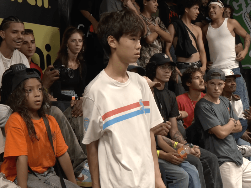 A young skateboarder in a white shirt with red and blue stripes stands focused in the center of a crowded indoor skate event, surrounded by spectators sitting and standing close together, some smiling and others watching intently.