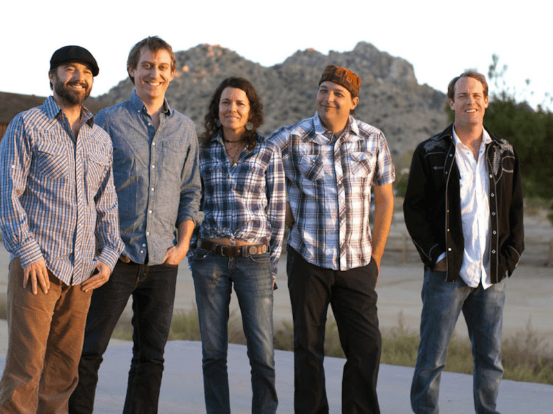 A group portrait of the five members of the band Donna the Buffalo standing outdoors in a desert landscape. From left to right: Kyle Spark, Mark Raudabaugh, Tara Nevins, Jeb Puryear, and David McCracken. They are all smiling and dressed in casual, roots-rock attire, including flannel shirts and denim. Tara Nevins, in the center, wears a blue plaid shirt and a decorative belt, while Jeb Puryear to her right wears a patterned hat. The background features a large, rocky hill under the soft glow of golden hour light, with a rustic wooden building visible on the left.