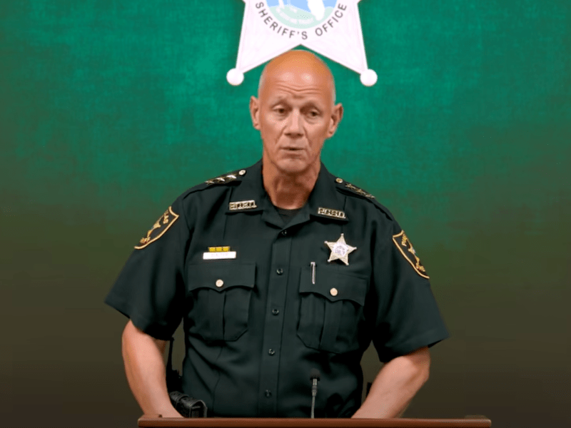 A bald man in a dark green sheriff's uniform, adorned with a star badge and shoulder epaulets, stands at a podium in front of a green backdrop with the Sheriff's Office seal visible above him.