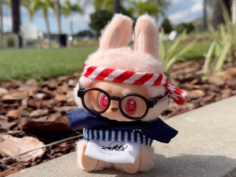 A fluffy, pink labubu toy is wearing a red and white striped headband, a dark blue shirt with a blue and white striped belt, a white apron saying "zukkus" and small round black glasses. It sits on a concrete ledge with mulch and green grass in the background, and palm trees in the distance under a bright sky.