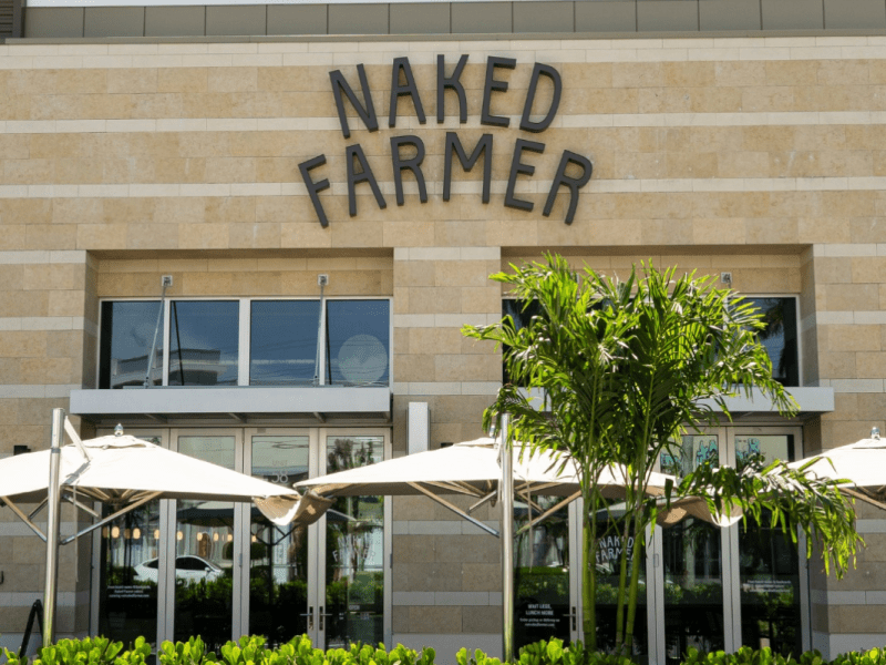 A straight-on, eye-level shot shows the exterior of a Naked Farmer restaurant. The building has a modern, tan-tiled facade with the name 'NAKED FARMER' in large, dark, sans-serif letters. In the foreground, an outdoor patio area features large white umbrellas, a small palm tree, and green bushes.