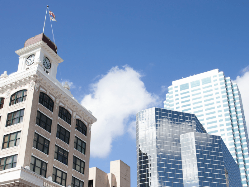 Low-angle view of the Tampa skyline, showing the historic City Hall building with its clock tower, contrasted against several taller, modern glass skyscrapers under a blue sky with white clouds.