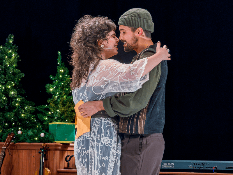 Two performers embracing on a dark stage lit by warm spotlights, likely from a holiday production. The person on the left has long, curly hair and wears a white lace overlay, while the person on the right wears a green knit beanie and a collared shirt. Small, illuminated Christmas trees stand on the set behind them.