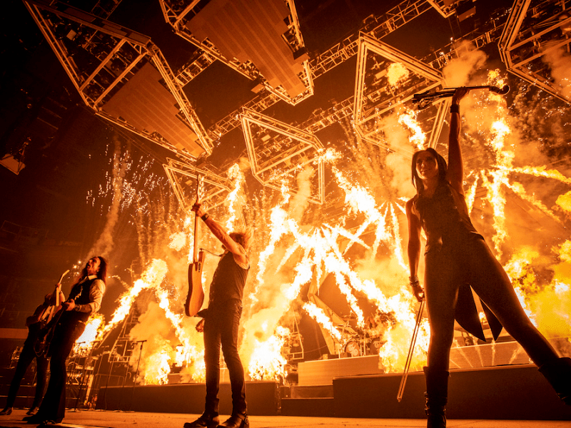 A highly dramatic concert photo showing the band in silhouette against bright orange and yellow flames. The stage rigging above is visible in the intense, high-contrast lighting.
